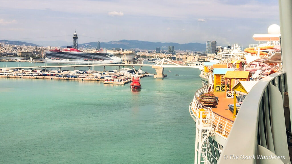 View of Virgin Voyages ship docked in the Port of Barcelona, seen from the upper deck of Oasis of the Seas with colorful cabanas and city skyline in the background.